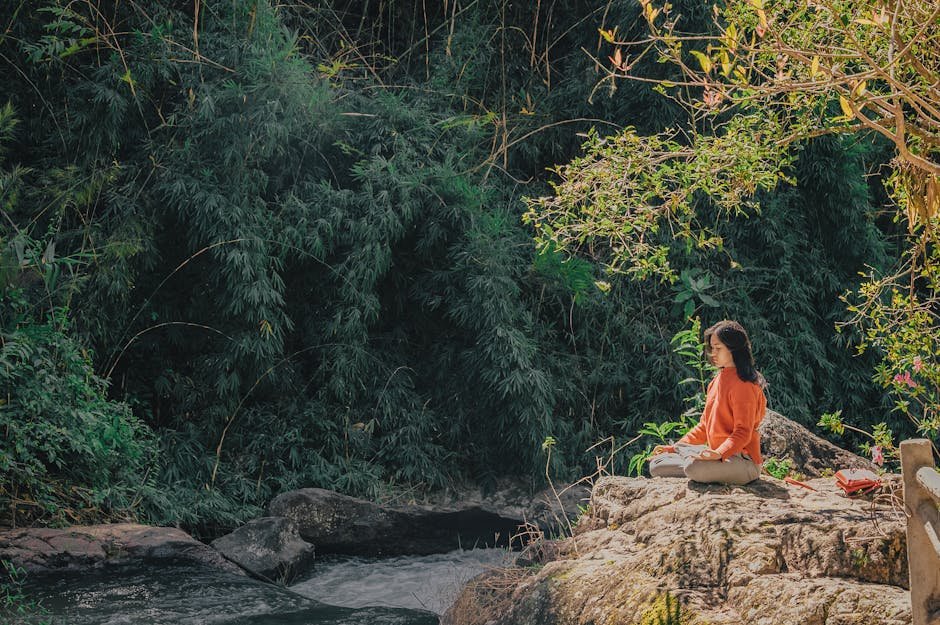 Woman meditating by a serene riverside surrounded by lush greenery on a sunny day.